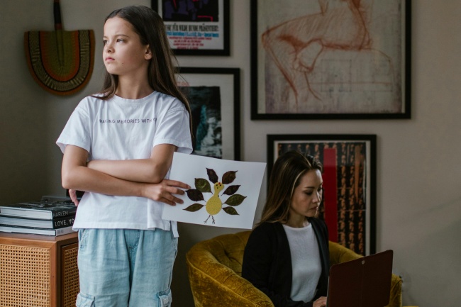 Ein Mädchen steht traurig mit einem Bild neben ihrer arbeitenden Mutter - die Muster von Eltern werden oft übernommen