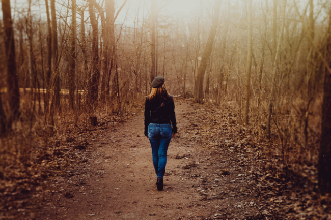 Zeit zu gehen: Eine Frau mit Jeans, dunkler Jacke und Mütze geht allein auf einem Waldweg zwischen kahlen Bäumen dem warmen Sonnenlicht entgegen – Symbol für Loslassen und Neuanfang.