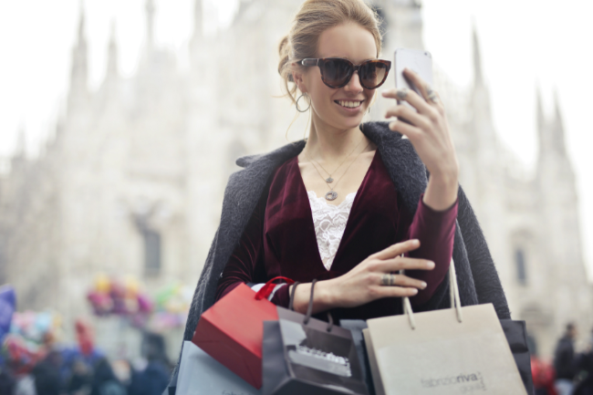 Frau mit mehreren Einkaufstüten schaut lächelnd auf ihr Smartphone – Symbol für Kompensationskauf und den kurzfristigen Glücksmoment beim Shoppen.